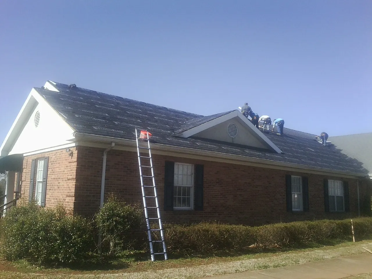 Crew tearing shingles off a brick ranch roof with a ladder set — Emergency Roof Repair work by our crew in Chataignier