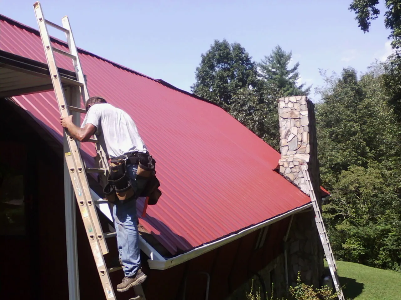 Roofer on a ladder beside a red metal roof and stone chimney — Soffit & Fascia Repair by Roofing Brothers in Chataignier
